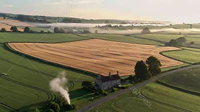 Aerial view of a rural house surrounded by vast golden wheat fields and green meadows in early morning mist