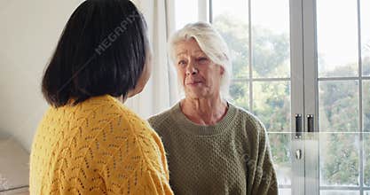 Diverse senior female friends standing by glass door speaking, wiping tears and sharing support