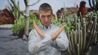 Man in striped shirt making an x gesture with arms in an outdoor park setting surrounded by cacti, expressing refusal or