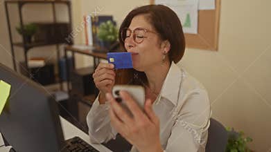 Woman holds creditcard while viewing smartphone at a computer desk in office beside corkboard and potted plant; happiness