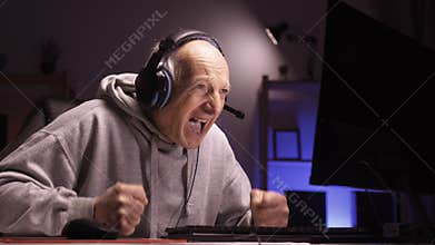Nervous senior mature gamer streaming his nighttime gaming session using personal computer bangs his fists on the table