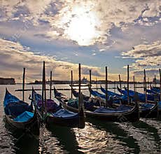 Moored Gondolas Adorn Venice Lagoon as Sunlight Breaks Through Clouds, Reflecting on Shimmering Waters, with Historic City in