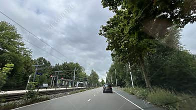 Tree lined urban road beside tram stop and car traffic under cloudy