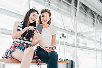 Asian girls using smartphone checking flight or online check-in at airport together, with luggage. Air travel, summer holiday