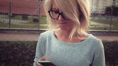 Sad young woman standing in the park area among buldings using her smartphone texting feeling sad