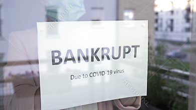 Woman in a medical mask hangs a BANKRUPT sign on the window. Impact of the COVID-19 pandemic on the small and medium