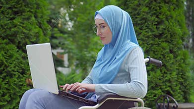 Portrait young arab woman disabled in a traditional scarf uses a laptop sitting in a wheelchair