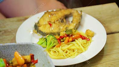Female hand puts vegetables in a plate with spaghetti and fish.