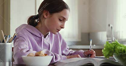 Smart adolescent schoolgirl doing homework alone in kitchen.