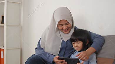 Asian muslim mom and little baby girl daughter learning online or watching videos on tablet phone, happiness between mother and