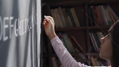 Indian school girl pupil holding chalk writing english letters on blackboard.