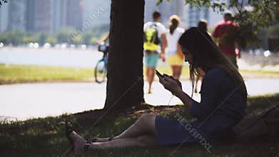 Young beautiful woman sitting on grass in park and using smartphone. Female resting in downtown of Chicago, USA.
