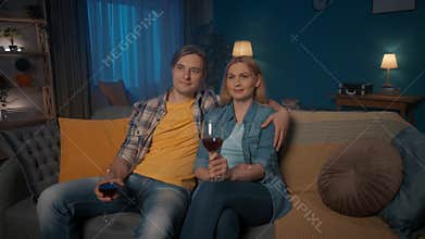 A young couple sits hugging on the couch with glasses of red wine and watching TV. A man and a woman are watching a