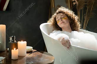 Pleasant caucasian woman puts on eye patches and relaxes in a bubble bath. The woman spends the evening relaxing in the