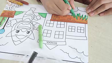 Coloring the green grassy area using dark green crayon. An Asian girl is coloring. View from above.