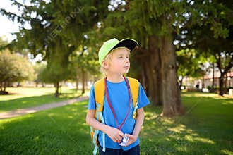 Elementary boy wearing baseball cap and with backpack and name badge on his neck goes to school. Child is waiting of school bus.