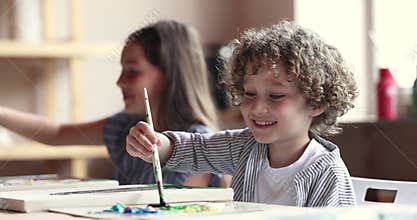 Little boy holding paintbrush painting on canvas seated at table