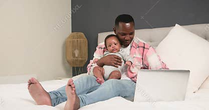 Father Balances Work and Family: A Loving Dad Works on His Laptop While Holding His Adorable Baby on the Bed
