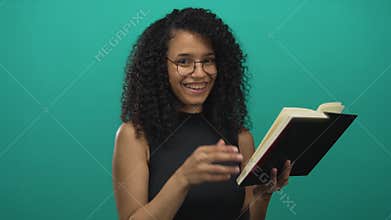 Young hispanic woman reading book over isolated green background wall looking amused and thoughtful