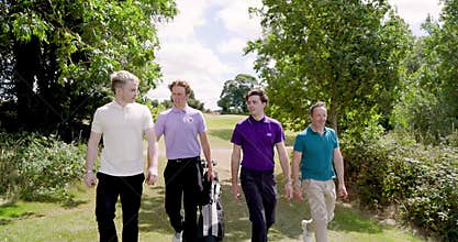 Male golf players wearing uniforms, walking with clubs and talking on golf course on sunny day