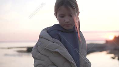 Beautiful little girl in warm jacket standing by the seashore at sunset