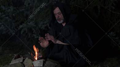 A mysterious hooded monk prays by the fire with a rosary.