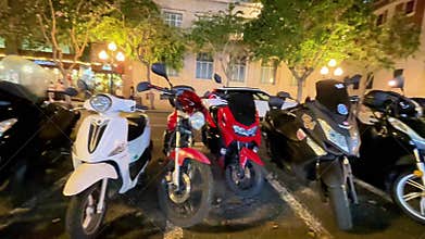 Scooters and motorcycles parked at night in a city street with trees and buildings