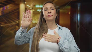 Woman in hotel taking oath with hand raised, showing commitment and honor, wearing a casual shirt, standing inside a warmly lit