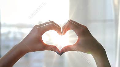 The girl in her bedroom at the window catches her hands, made in the form of a heart, the sun`s rays. Bokeh