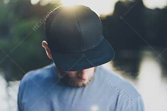 Photo Bearded Young Man Wearing Black Blank Cap. Green City Park Lake Background and Sunset effect. Front view