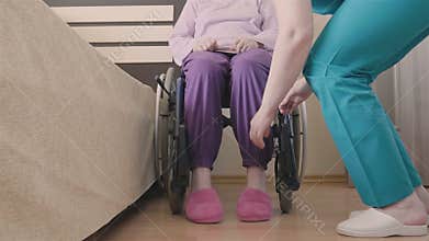 Young nurse caring of elder disabled woman in wheelchair and transfers her in bed