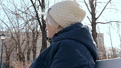 Portrait of a lonely depressed elderly senior woman sitting on a bench in city park in early spring in sunny day -