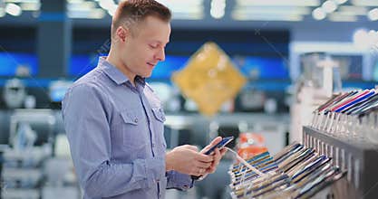 A young handsome man standing at a shop window with smartphones in a portable electronics store chooses to buy a new