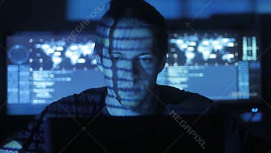 Male hacker programmer working at computer while blue code characters reflect on his face in cyber security center