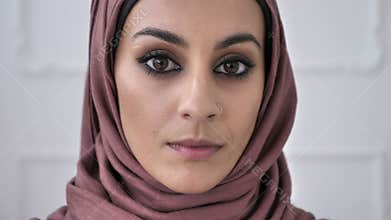 Young beautiful serious Indian girl with piercing in the nose in pink hijab looking at camera. Portrait, tree, white