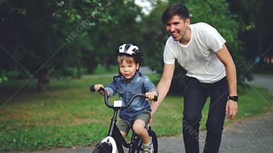 Slow motion of laughing child cycling in park with careful father who is teaching him to ride bicycle. Happy young