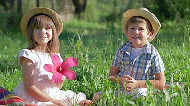 Little couple in love outdoor, boy gives pink flower to pretty girl sitting in grass