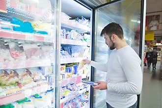 A man chooses frozen foods from shelves in a refrigerator in a supermarket. A man buys products in the store