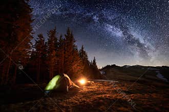 Night camping. Illuminated tent and campfire near forest under night sky full of stars and milky way