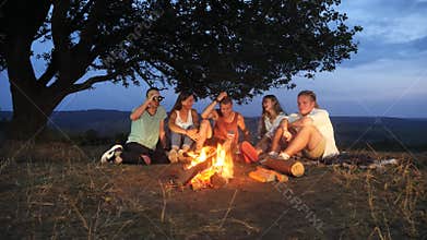 Group of friends sit next to a campfire with warm drinks and talk