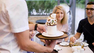 Hospitable waiter walking to the table bringing tasty dessert with whipped cream and chocolate for little client. Happy