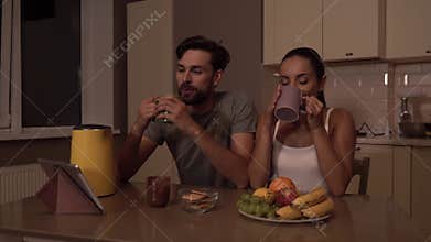 Guy is sitting down in chair and looking at girl. She looks at him too and drinks from cup. They are smiling to each