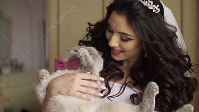 Portrait of the young adorable bride with charming smile and long dark curly hair playing with lovely cat with pink bow