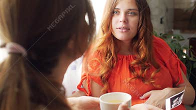 Two young women sitting in kitchen with cup of tea and discussing happy pregnancy of one, looking at an ultrasound scan, sisters c