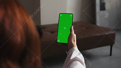 Caucasian young woman holds phone, with empty green screen in vertical mode. App advertising. No touch, talking head