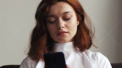 Beautiful caucasian woman sitting on a blue sofa and using smartphone or cell phone in modern room. Tapping, scrolling