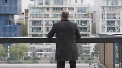A man is standing on a balcony, looking out at the cityscape in an urban setting