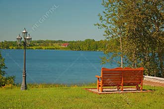 A park bench and a lamppost on the grass facing a blue lake under a blue sky