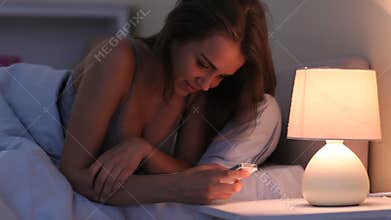 Pretty smiling brunette lying in bed texting on smartphone