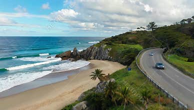 Coastal road winds along sandy beach with turquoise ocean and rocky cliffs under a cloudy sky